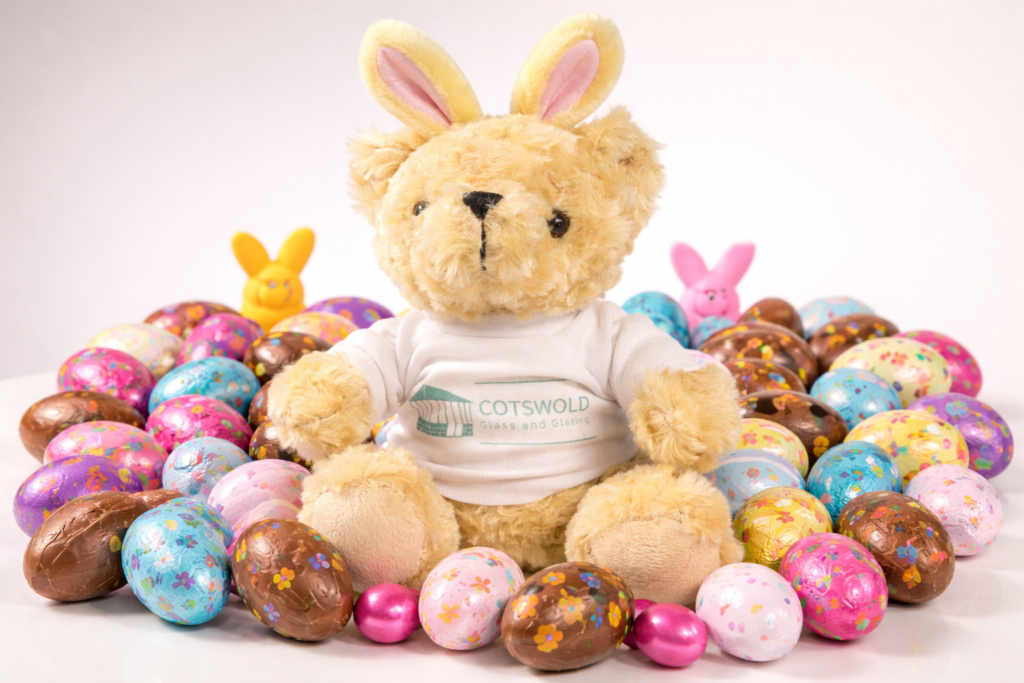 Cotswold Glass teddy bear with bunny ears surrounded by colourful Easter eggs.